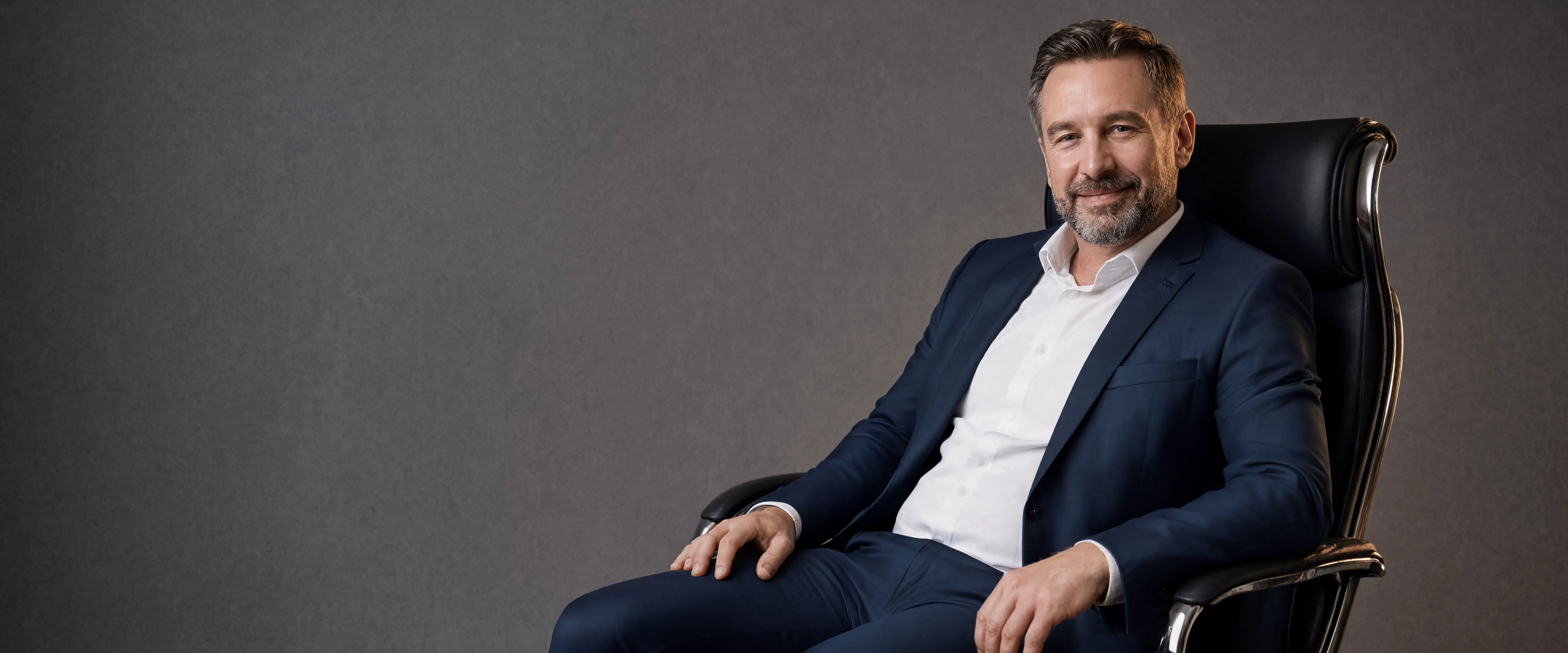 A man with a beard wearing a navy blue suit jacket and white shirt, sitting in a black leather office chair against a dark gray studio background.